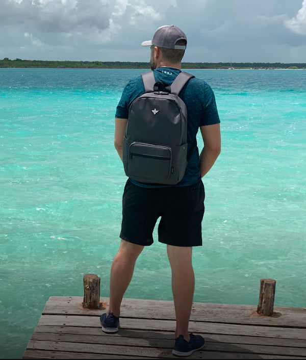 A man with a backpack stands on a wooden dock, facing a turquoise body of water under a cloudy sky.