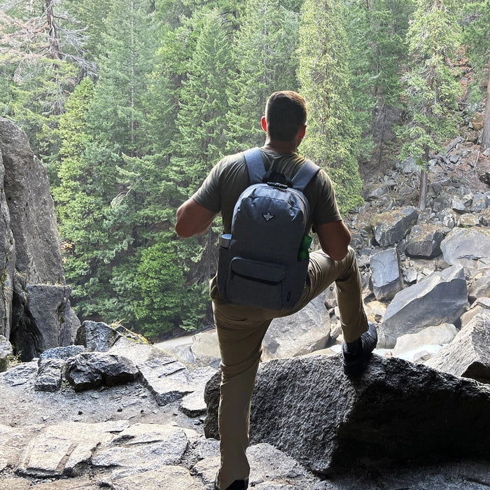 Person with a backpack stands on a rocky ledge, looking out at a forested area with large rocks and trees in the background.