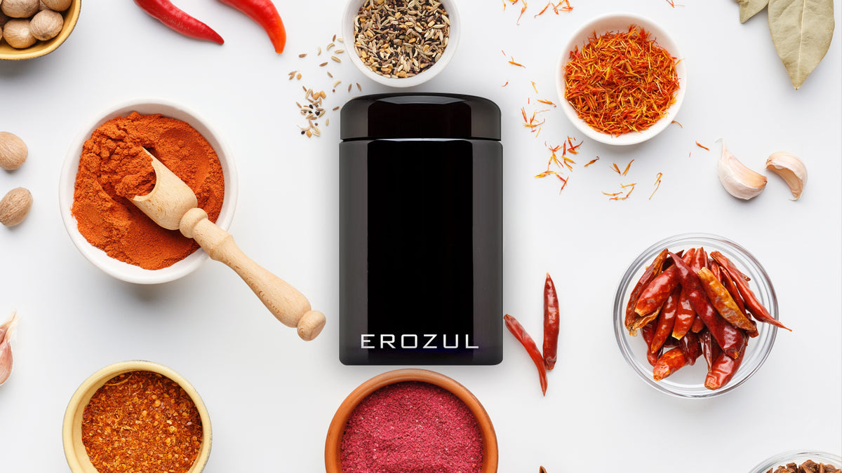 A black Erozul container is surrounded by various spices, including dried chilies, saffron, and ground powders in bowls, arranged on a white surface.