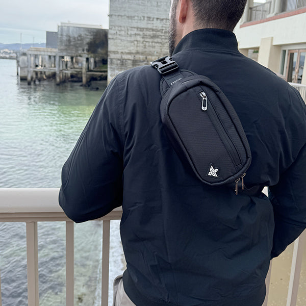 A person in a black jacket with an Erozul Fortuna Smell Proof Crossbody Fanny Pack Bag stands at a railing overlooking water and industrial buildings.