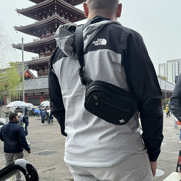 A person in a black and gray jacket carries the Erozul Fortuna Smell Proof Crossbody Fanny Pack Bag while facing a Japanese pagoda, with people walking in the background.
