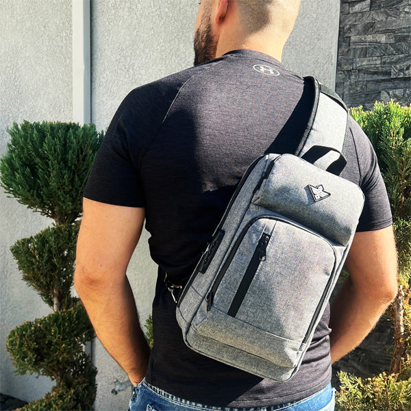 A man stands outdoors near trimmed bushes and a stone wall, wearing the Erozul Equinox Smell Proof Sling Bag With Combination Lock over his right shoulder.